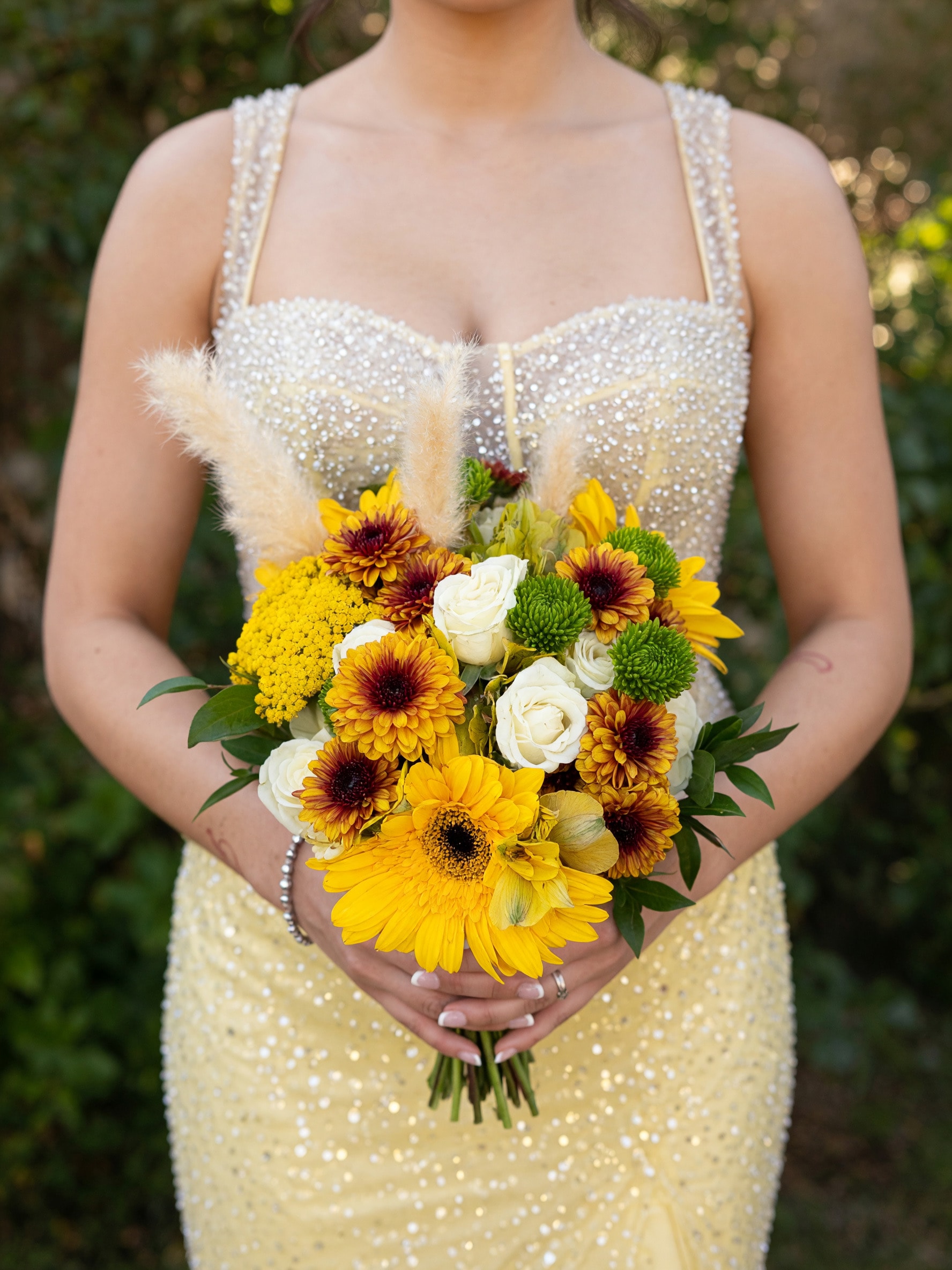 Bride in beaded gown holding yellow sunflower gerbera and white rose bouquet with pampas grass outdoors