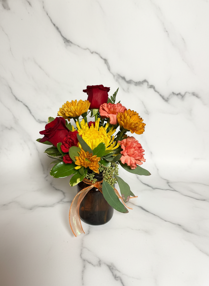 Fall arrangement with red roses, yellow and orange mums, and coral carnations in amber mason jar with ribbon on marble surface
