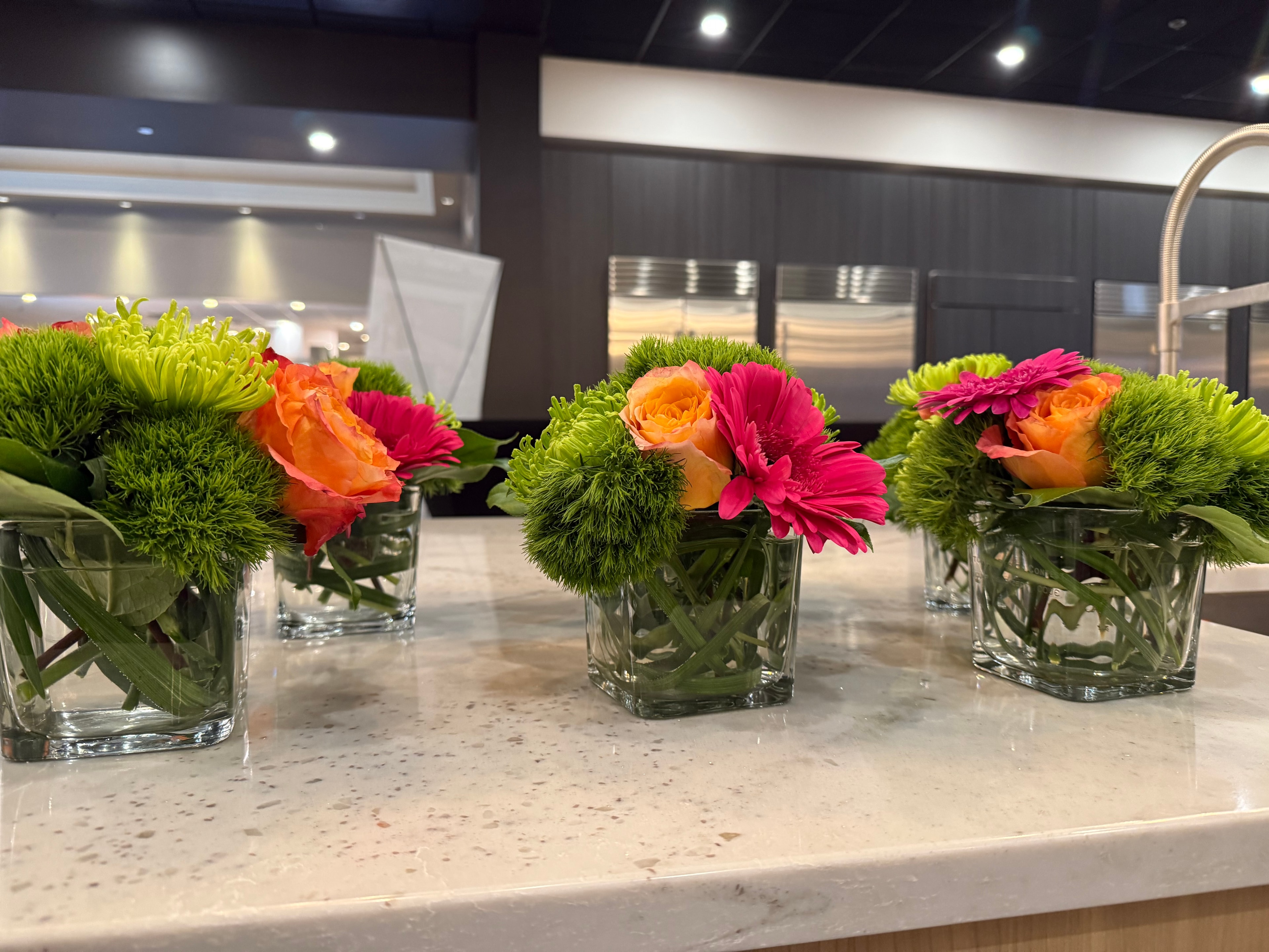 Three square glass cube arrangements with orange roses, hot pink gerberas, and lime green chrysanthemums on marble counter