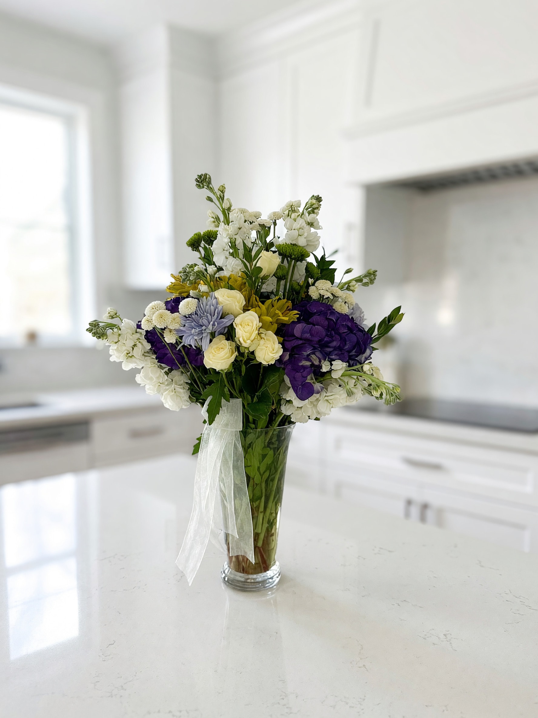 Purple hydrangeas, white roses, and mixed blooms in a tall glass vase on a kitchen counter