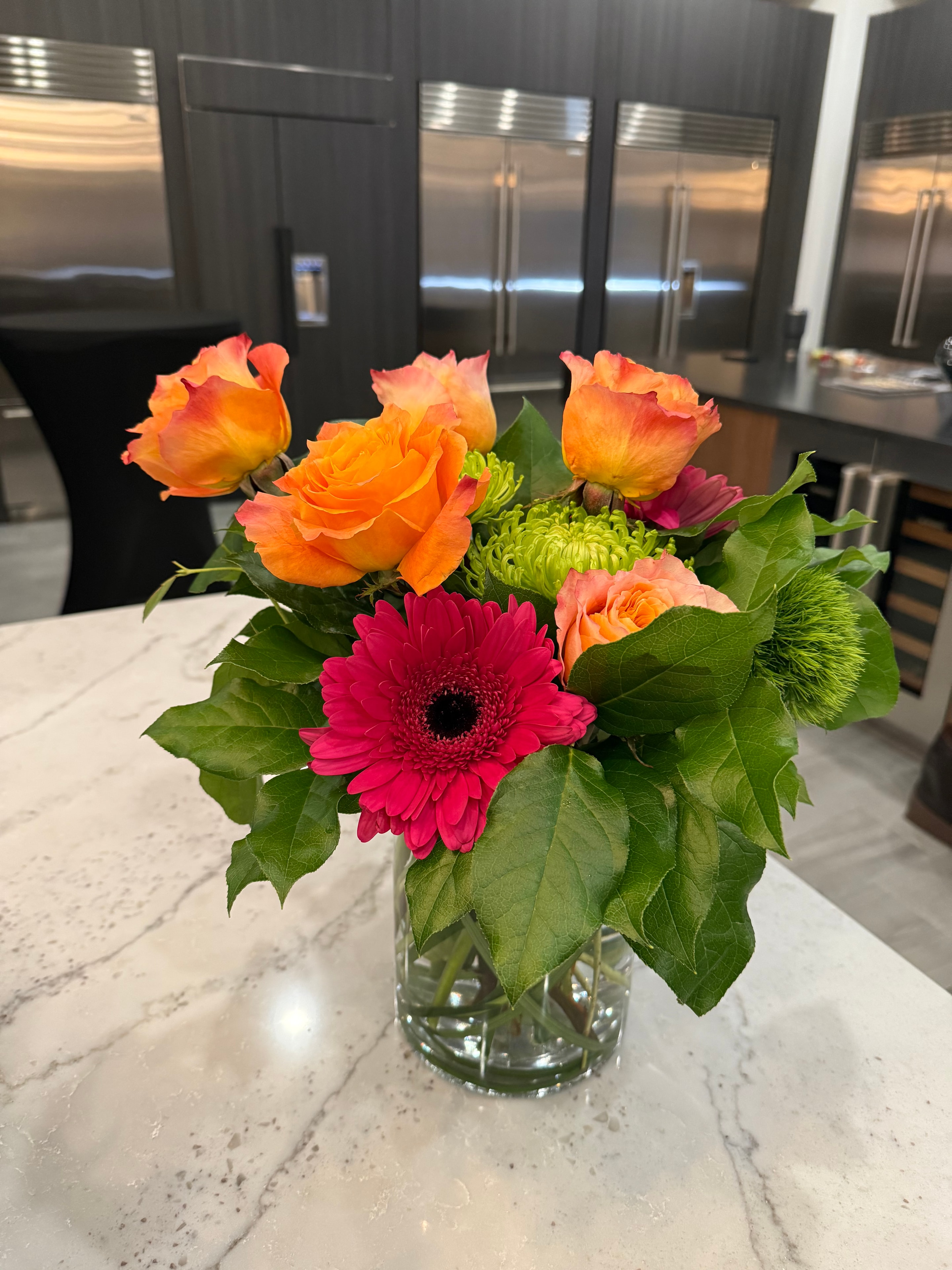 Orange roses and pink gerberas with green foliage in a glass vase on marble counter